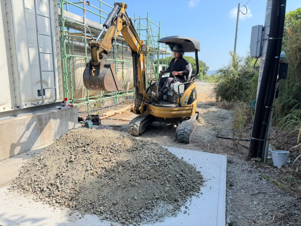 （重機での作業）: 種子島の受水槽基礎工事現場で重機を用いて砕石整地作業を行う様子