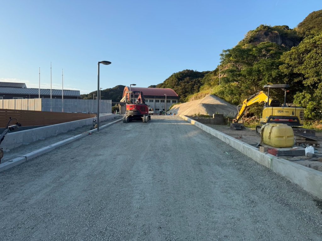 <img src="image_4.png" alt="体育館へと続く、綺麗に整地・砕石敷きされた進入路と土木工事用の重機">